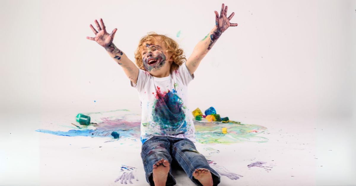 An enthusiastic child sitting on the floor, covered in paint, reaching their arms up and laughing.